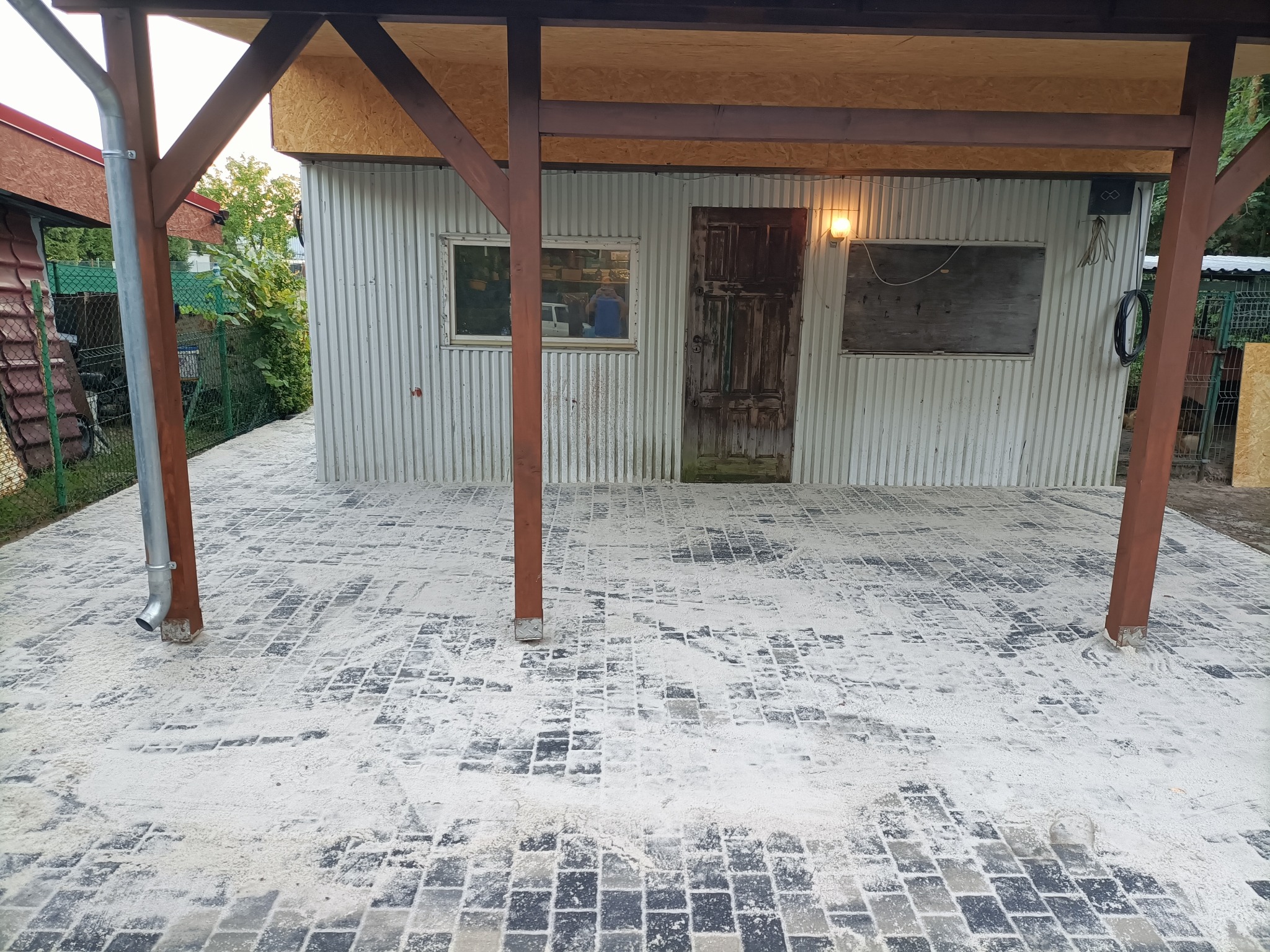 Świeżo ułożona, czarno-szara kostka brukowa pod zadaszeniem, posypana piaskiem. Widoczne metalowe ściany budynku i drewniane podpory dachu.