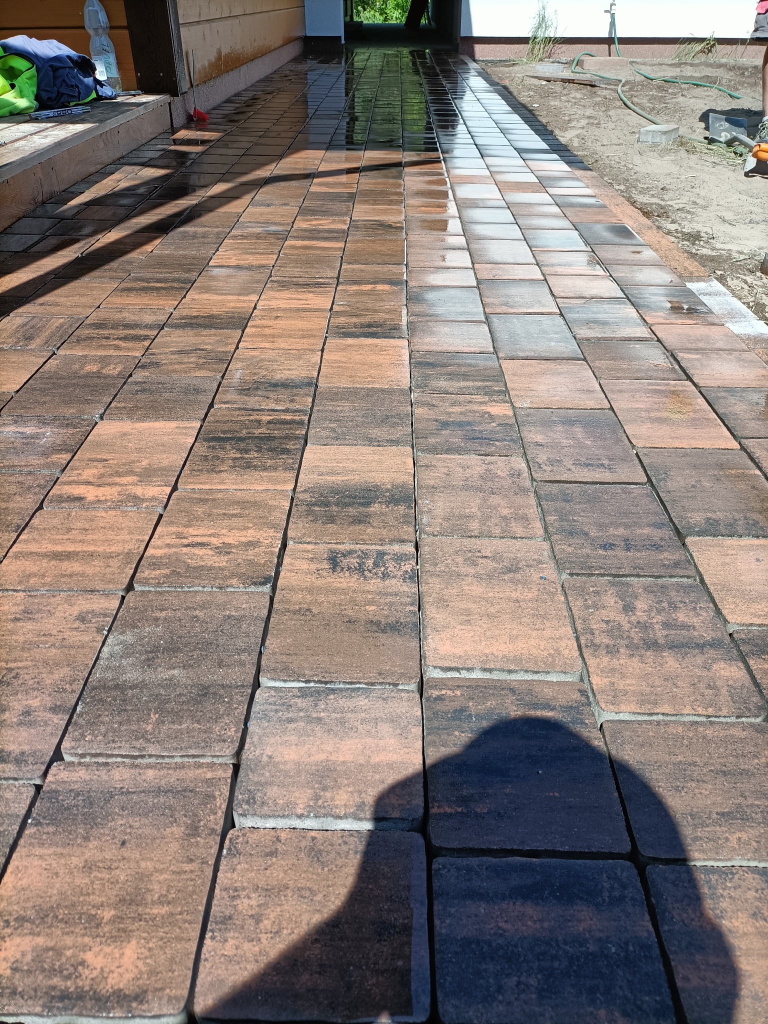 Świeżo ułożona, mokra kostka brukowa w odcieniach brązu i szarości, tworząca ścieżkę. Widoczne narzędzia i materiały budowlane w tle, sugerujące trwające prace wykończeniowe.