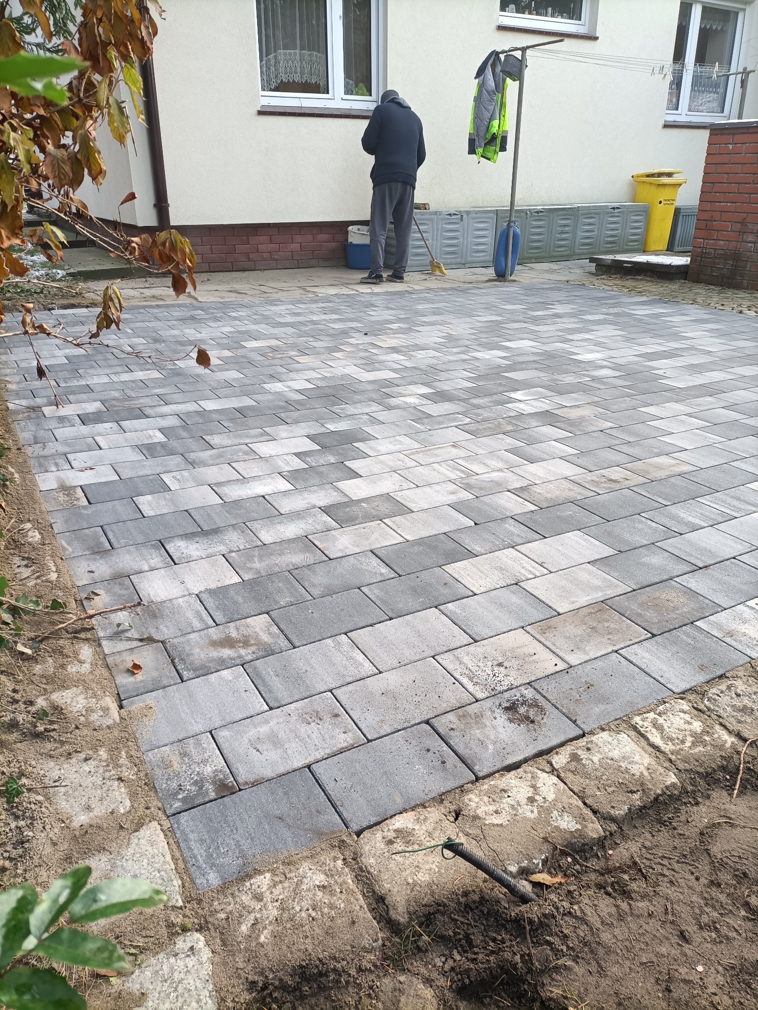 Ułożona szara kostka brukowa na posesji, z widocznym fragmentem domu i osobą zamiatającą. Krawężnik z jasnej kostki. Częściowo widoczne drzewo z brązowymi liśćmi.
