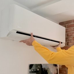 Sebastian kalinowski - Biały klimatyzator Midea na ścianie z cegły, montowany przez osobę w żółtej bluzie. Widoczny fragment obrazu pod klimatyzatorem.