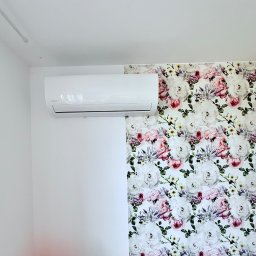Sebastian kalinowski - Biały klimatyzator na ścianie, z lewej biała ściana, z prawej tapeta w kwiaty. Widok z dołu, urządzenie zamontowane blisko sufitu.