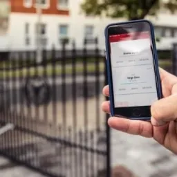 Otwarta brama wjazdowa sterowana smartfonem z aplikacją do automatyki domowej, widoczna ręka trzymająca telefon.