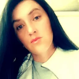 Portret kobiety z ciemnymi włosami i jasną cerą, skierowanej lekko w lewo, z widocznym fragmentem ubrania i rozmytym tłem z obrazami.