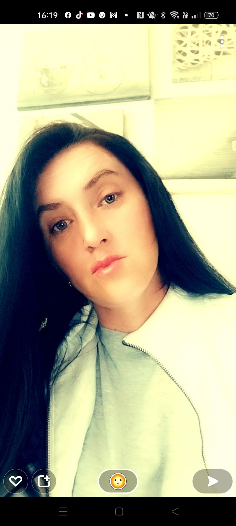 Portret kobiety z ciemnymi włosami i jasną cerą, skierowanej lekko w lewo, z widocznym fragmentem ubrania i rozmytym tłem z obrazami.