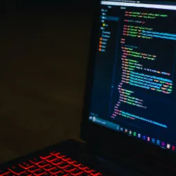 Ekran laptopa z edytorem kodu, podświetlana na czerwono klawiatura, widoczne ikony popularnych aplikacji na dolnym pasku.