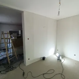Pokój w trakcie prac wykończeniowych: świeżo pomalowane ściany na biało, widoczne ślady po szpachlowaniu, drabina malarska i reflektory budowlane na podłodze.