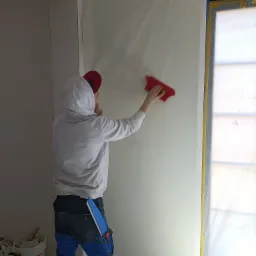 Pracownik w bluzie z kapturem wygładza szpachlą ścianę zabezpieczoną folią malarską, widoczne narzędzia w kieszeni spodni roboczych i wiaderka z zaprawą w tle.