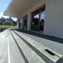 Wykończone schody zewnętrzne z szarego, ziarnistego materiału prowadzące do tarasu nowoczesnego domu, z widocznym pędzlem na krawędzi stopnia i pracownikiem w tle.