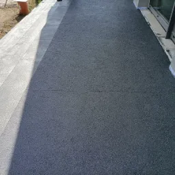 Nowoczesny taras z szarą, gumową nawierzchnią z widocznymi drobnymi granulkami, przylegający do budynku z szarą elewacją i dużym oknem tarasowym, obok ułożony chodnik z jasnoszarych płyt.