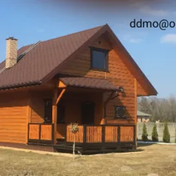 Gotowy, drewniany dom z brązowym dachem i ceglanym kominem, widoczny z zewnątrz, z małym tarasem i ogrodzeniem w tle, w słoneczny dzień.