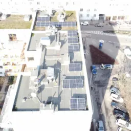 Widok z góry na instalację paneli na dachu budynku mieszkalnego, częściowo zacienione panele w trakcie montażu, obok widoczne narzędzia i przewody, w tle zabudowa miejska i ulica z samochodami.