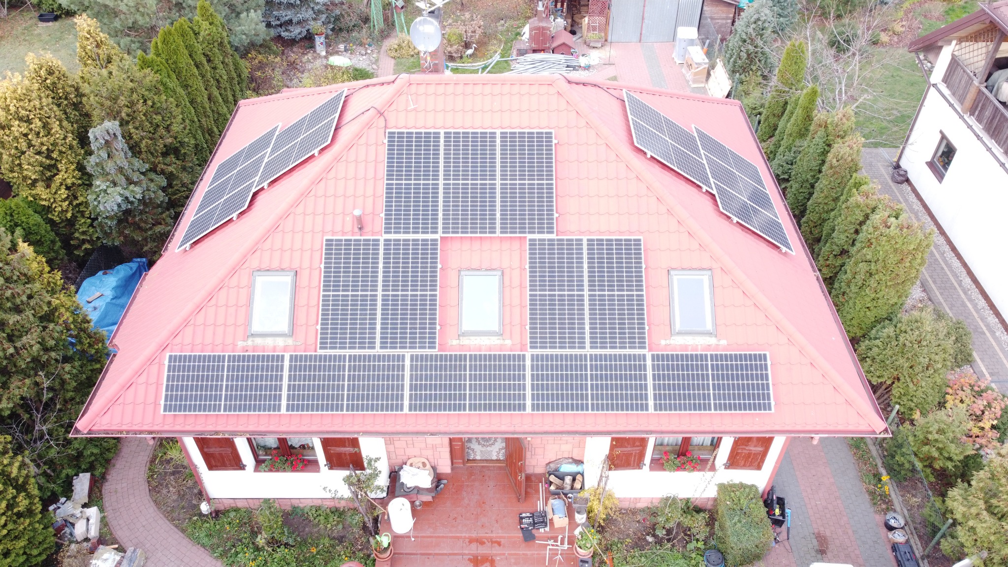 Widok z drona na dom w Zabrodziu z czerwonym dachem pokrytym panelami słonecznymi, otoczony zielenią i drzewami.