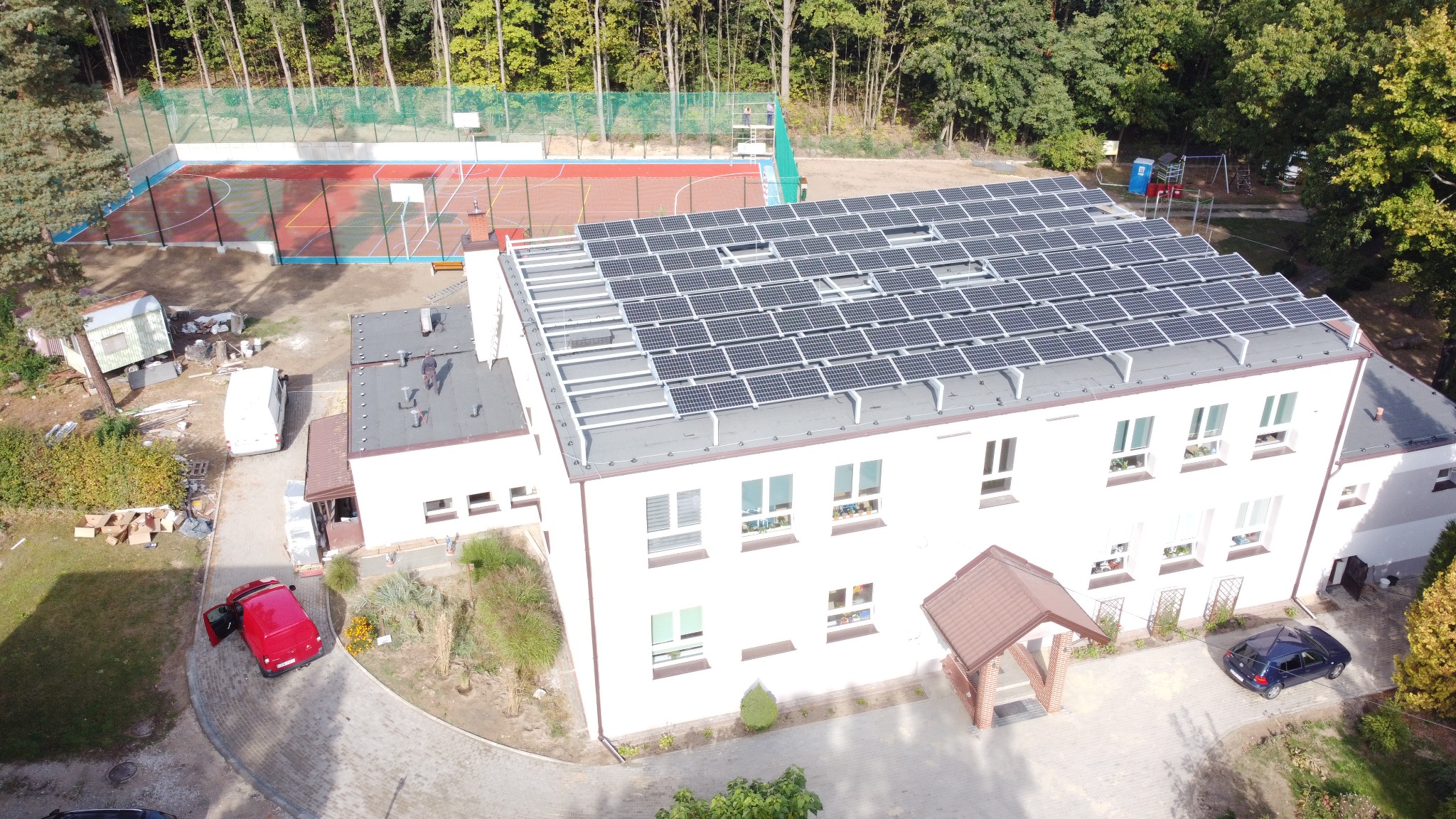Widok z góry na biały budynek z panelami słonecznymi na dachu, obok boisko sportowe, parking z samochodami i zieleń.