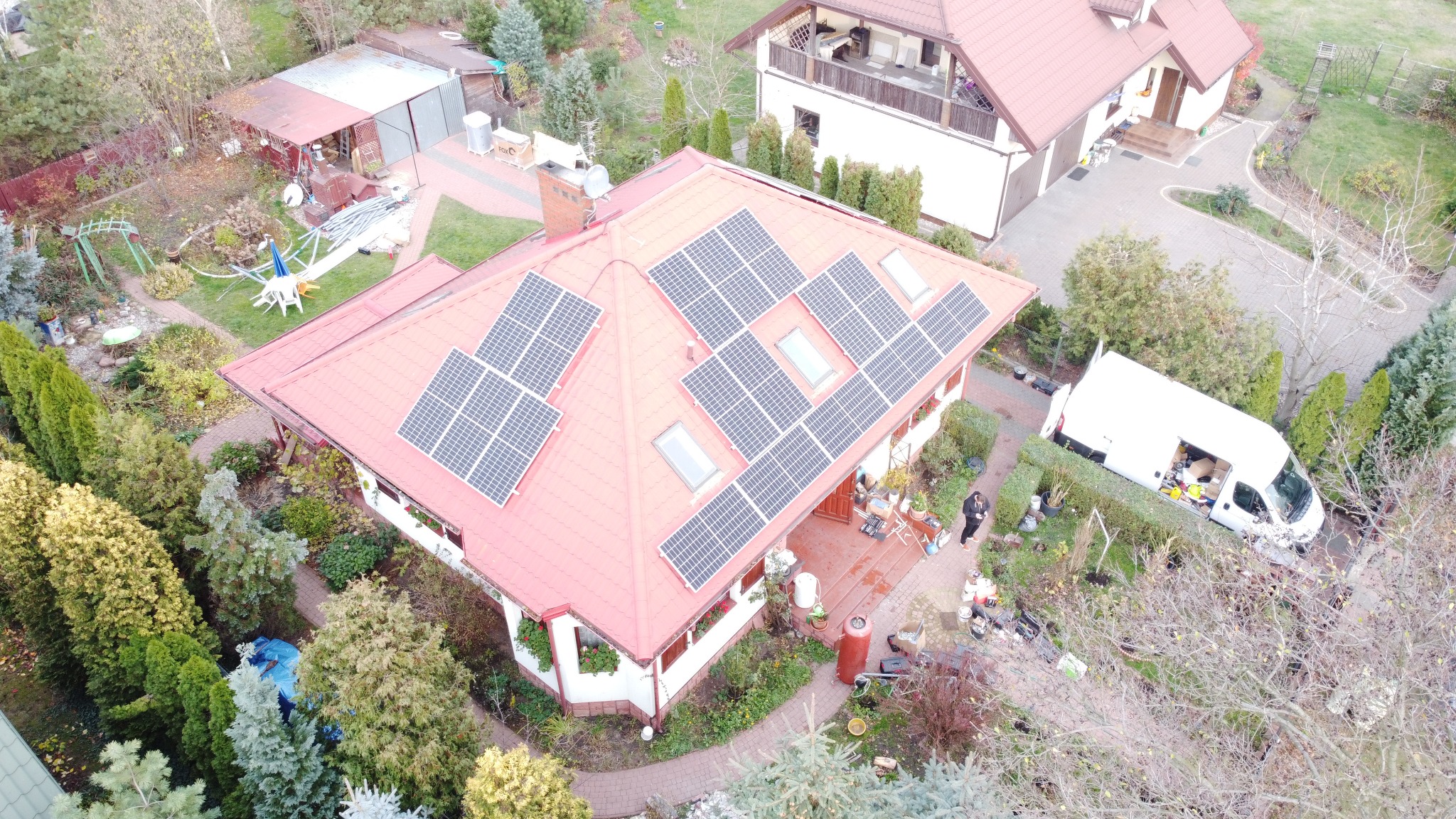 Widok z góry na dom z czerwonym dachem, na którym zamontowano panele słoneczne. Na podwórku stoi biały van z otwartymi drzwiami, a obok domu widać osobę.