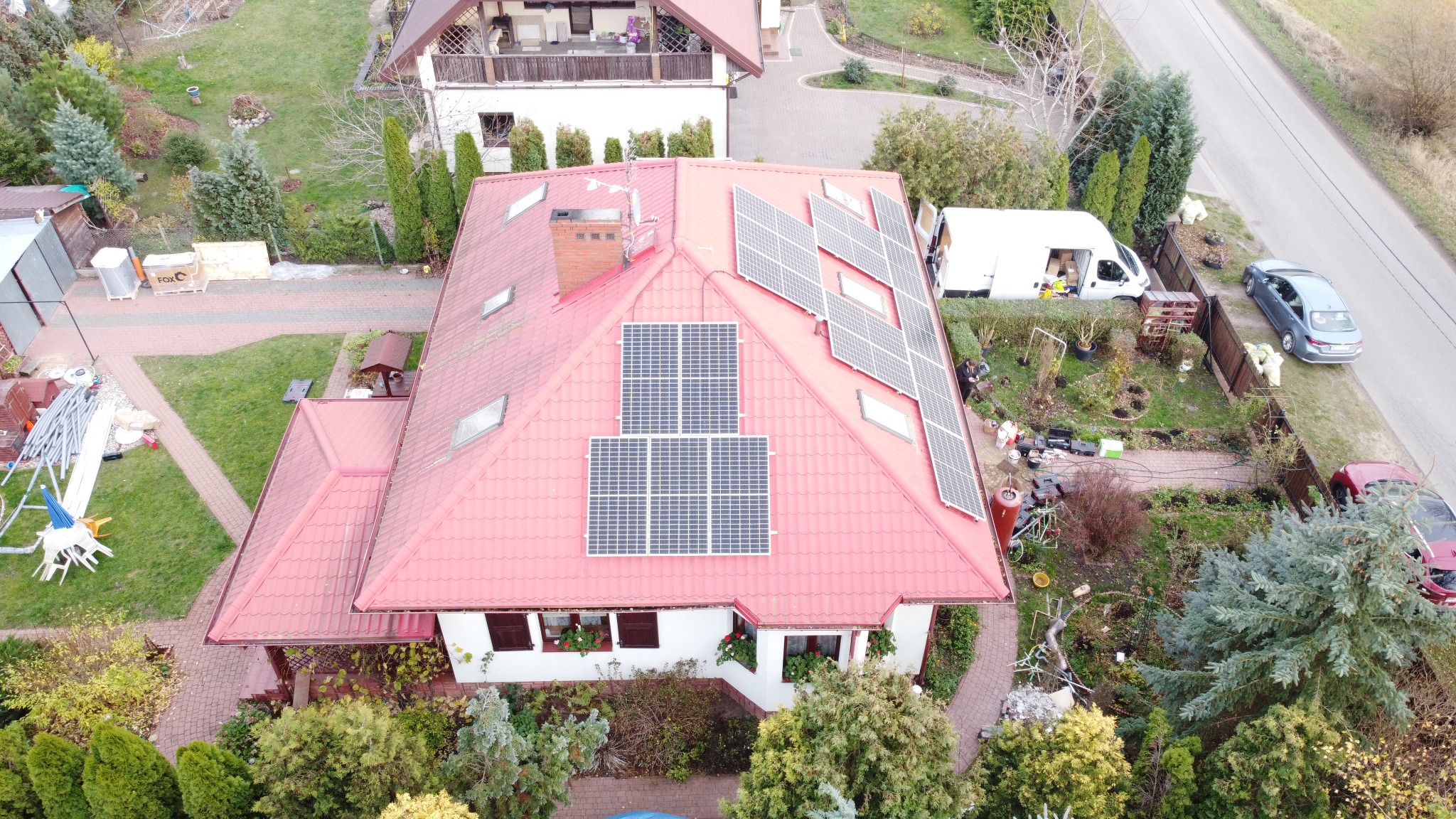 Widok z drona na dom z czerwoną dachówką, na której zamontowane są panele słoneczne. Na podwórku stoi biały samochód dostawczy i szary samochód osobowy. Widoczne narzędzia i materiały budowlane...