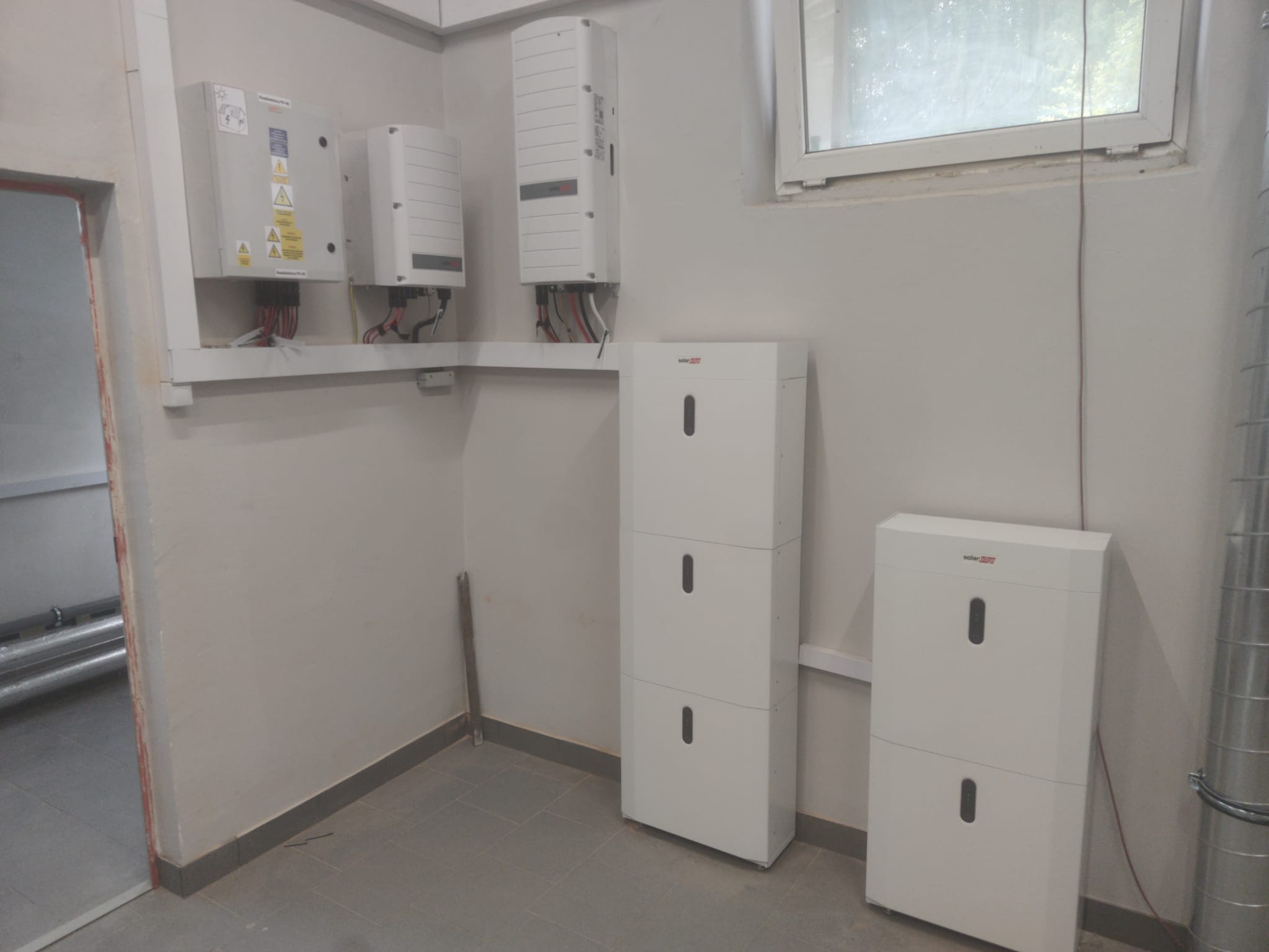Wewnętrzna instalacja z trzema białymi inwerterami na ścianie i dwoma modułowymi bateriami pod nimi, widoczne kable i rury instalacyjne przy ścianie, szara podłoga z płytek.