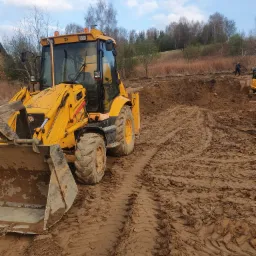 Żółta koparko-ładowarka JCB na placu budowy, widoczne ślady opon w błocie, w tle wykopany teren i sylwetka człowieka.