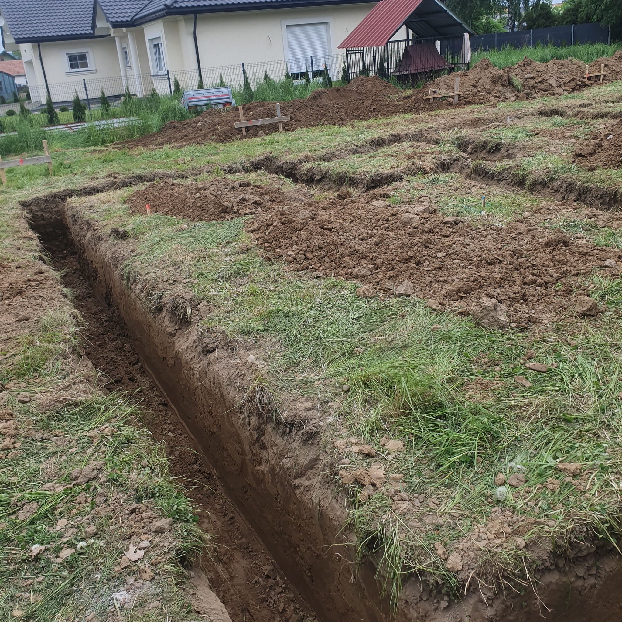 Wykopane rowy pod fundamenty domu jednorodzinnego, widoczne drewniane znaczniki, ziemia wymieszana z trawą.