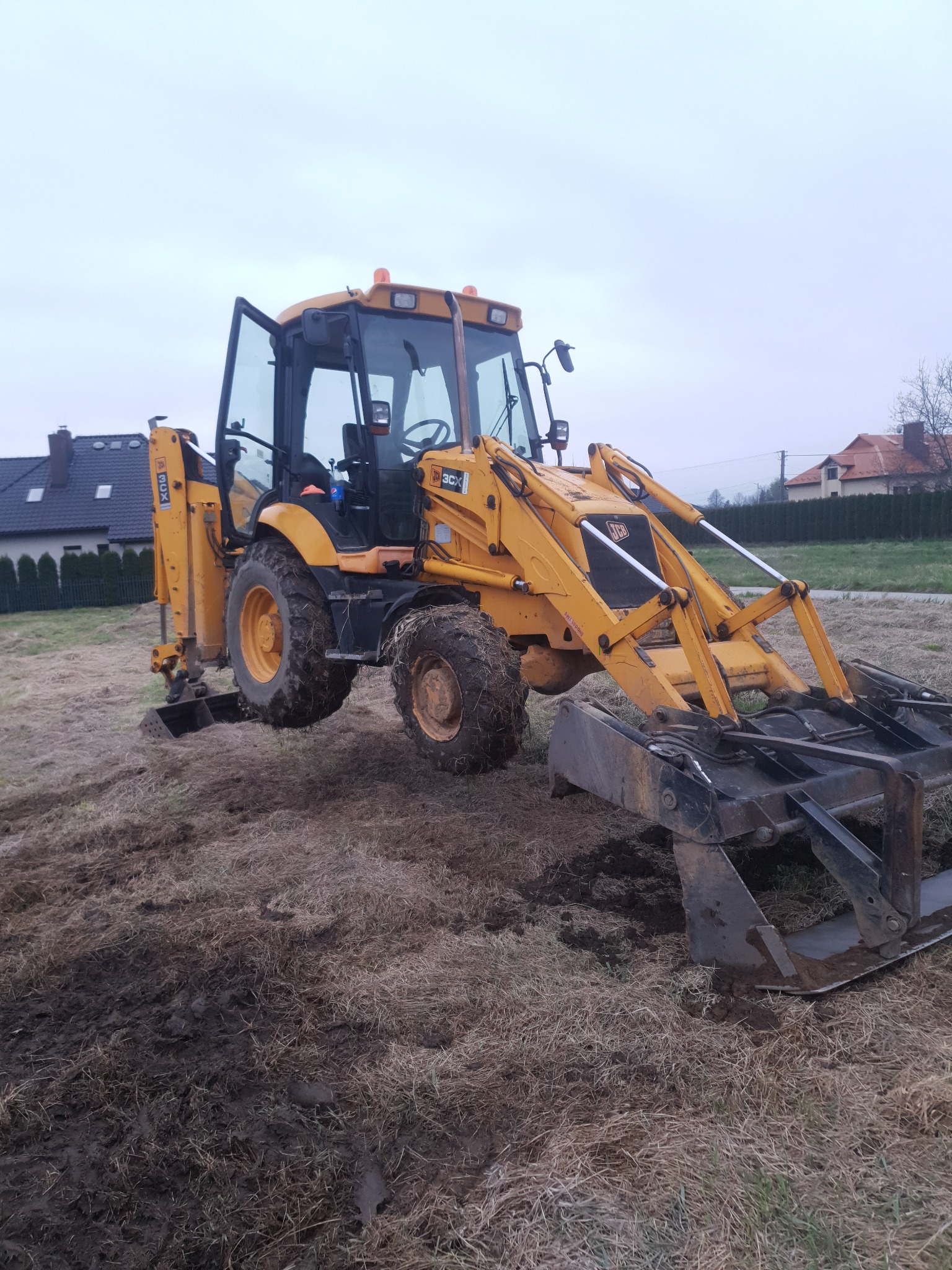 Żółta koparko-ładowarka JCB 3CX z otwartą kabiną stoi na nierównym, częściowo błotnistym terenie. W tle widoczne domy jednorodzinne pod pochmurnym niebem.