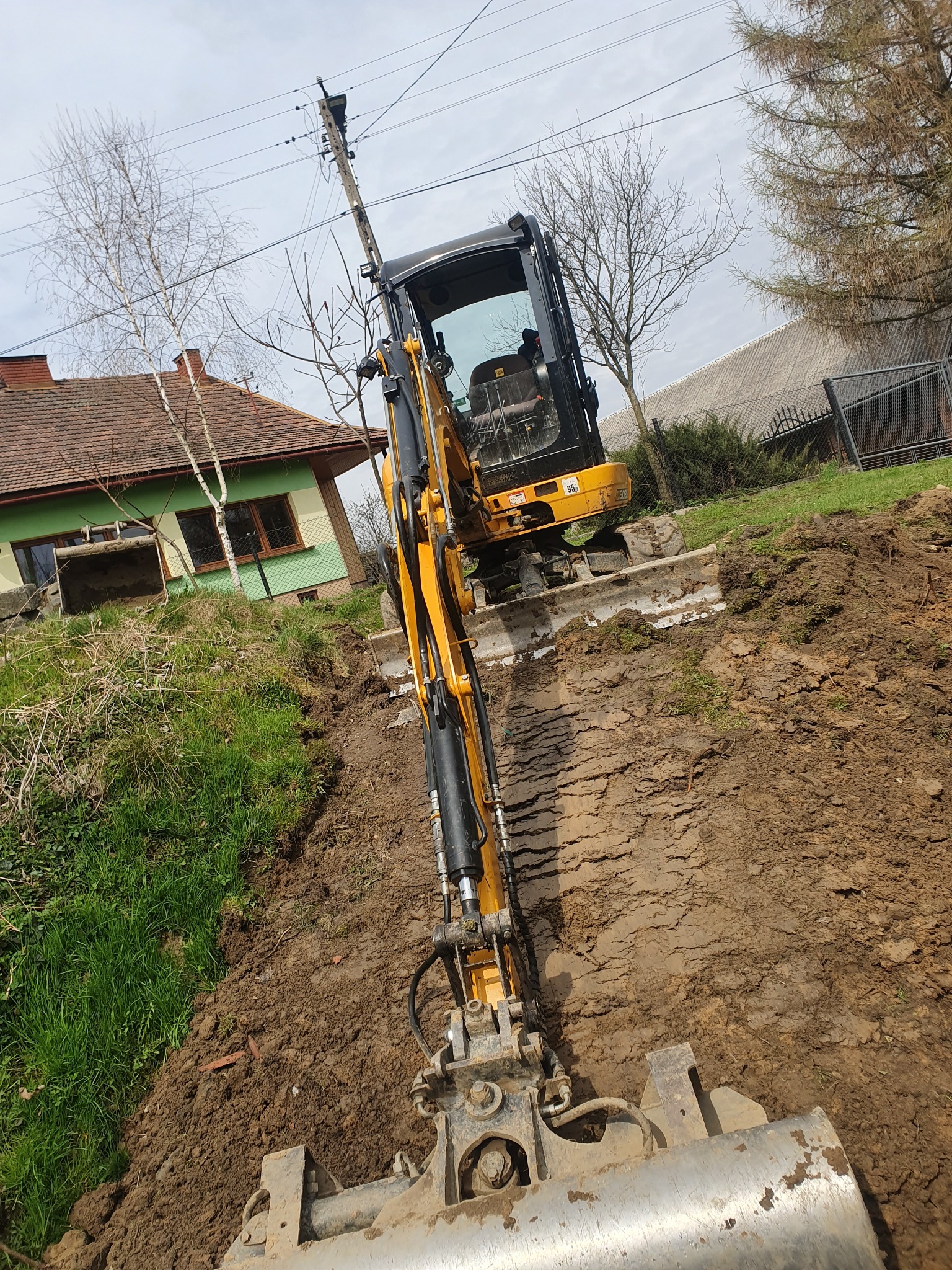 Żółta koparka gąsienicowa w trakcie prac ziemnych, widok z perspektywy łyżki, w tle zielony trawnik i dom z brązowym dachem.