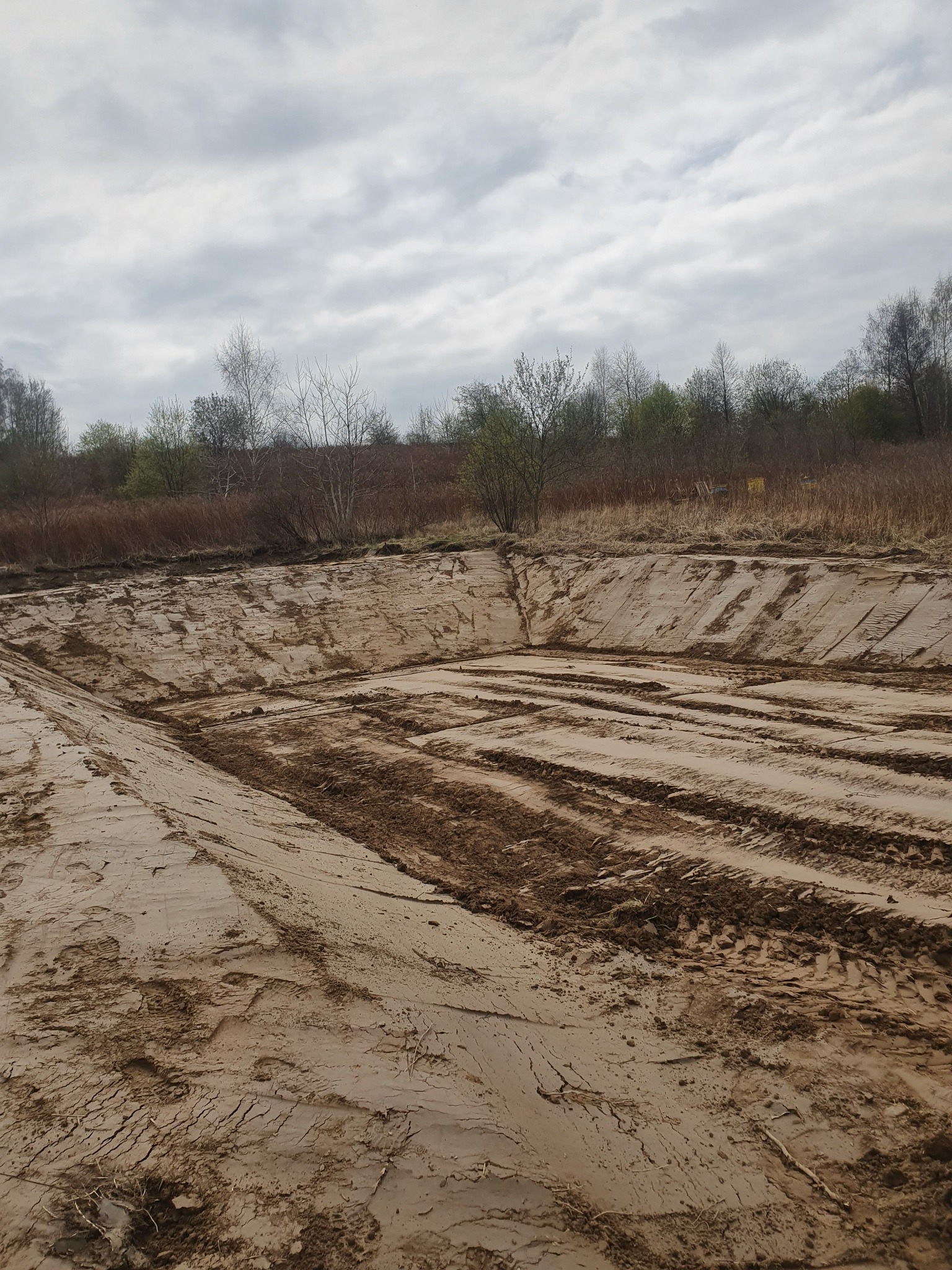 Wyrobisko w gruncie z widocznymi śladami opon, przygotowane pod budowę, w tle drzewa i pochmurne niebo.