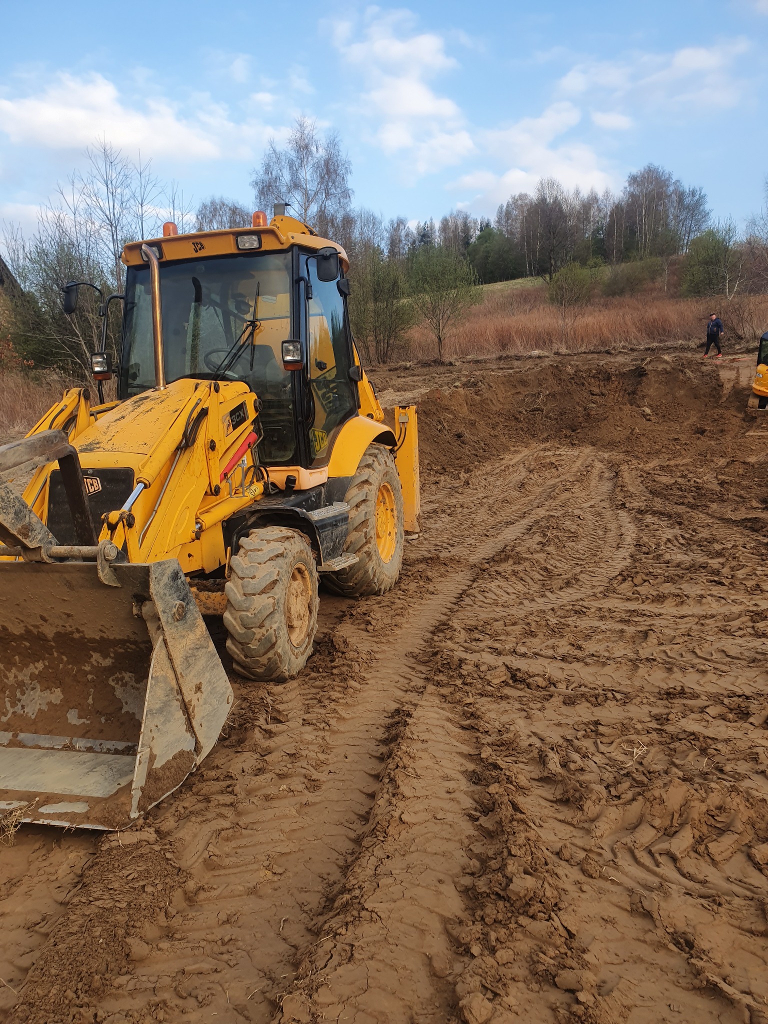 Żółta koparko-ładowarka JCB na placu budowy, widoczne ślady opon w błocie, w tle wykopany teren i sylwetka człowieka.