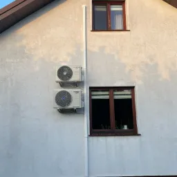 Dwa klimatyzatory marki Kaisai zamontowane na elewacji domu, widoczne okna z brązowymi ramami, biała rynna pionowa, jasna elewacja z delikatną fakturą.