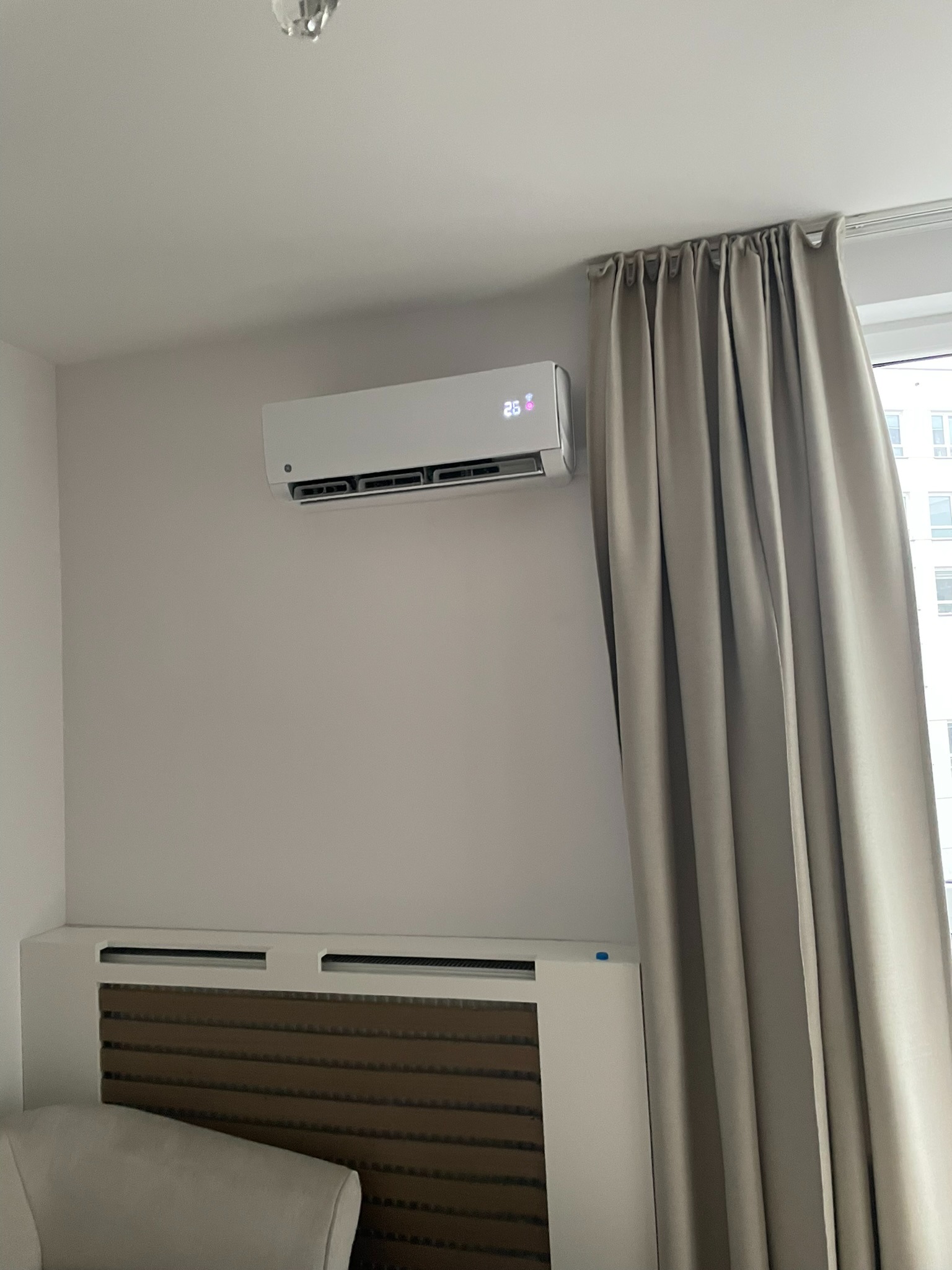 Biały klimatyzator ścienny z wyświetlaną temperaturą 26 stopni Celsjusza, zamontowany nad zabudowanym grzejnikiem w pokoju z beżowymi zasłonami.