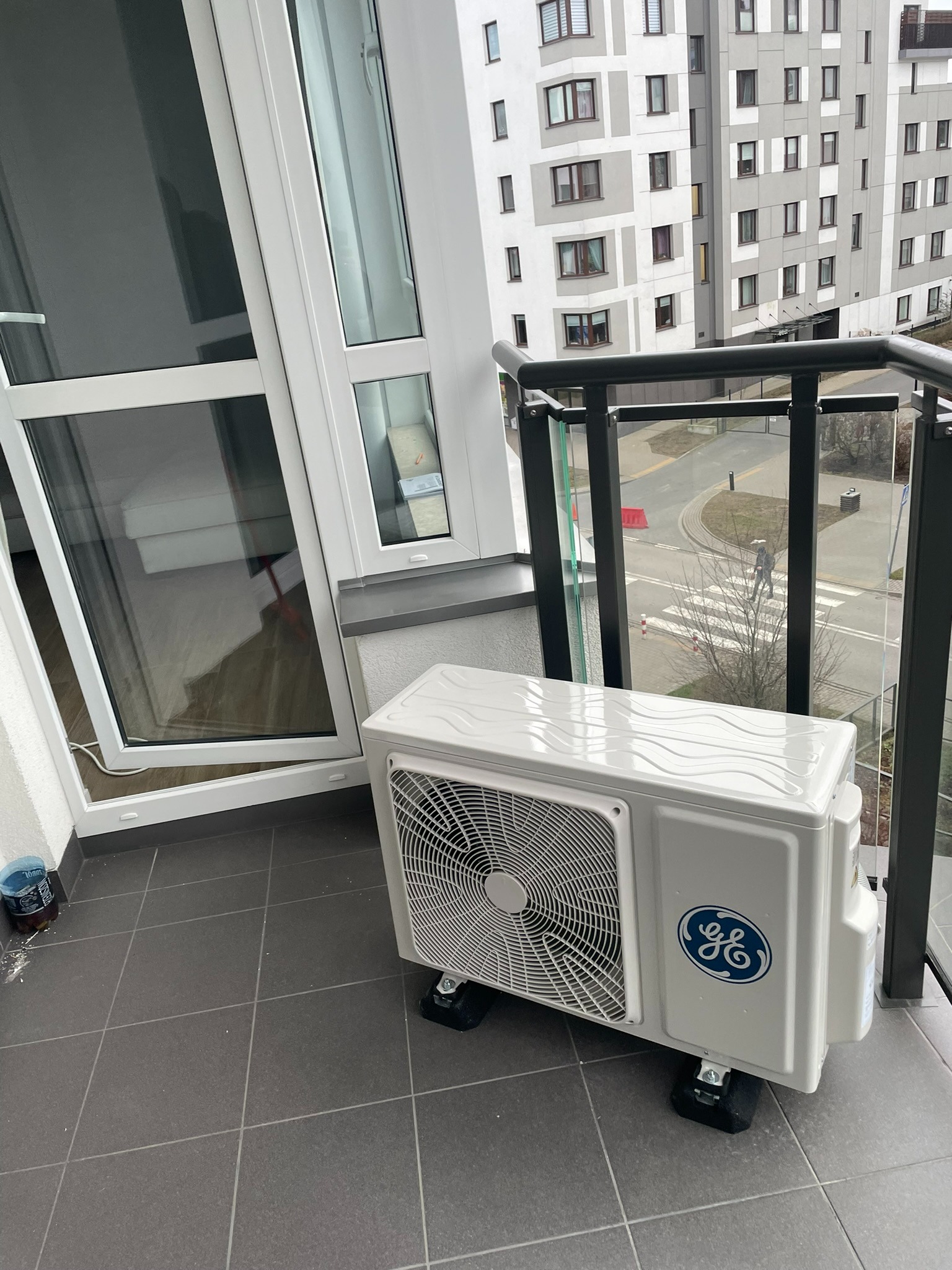 Biała jednostka zewnętrzna klimatyzacji z logo GE na balkonie, widok na osiedle mieszkaniowe i otwarte okno balkonowe.