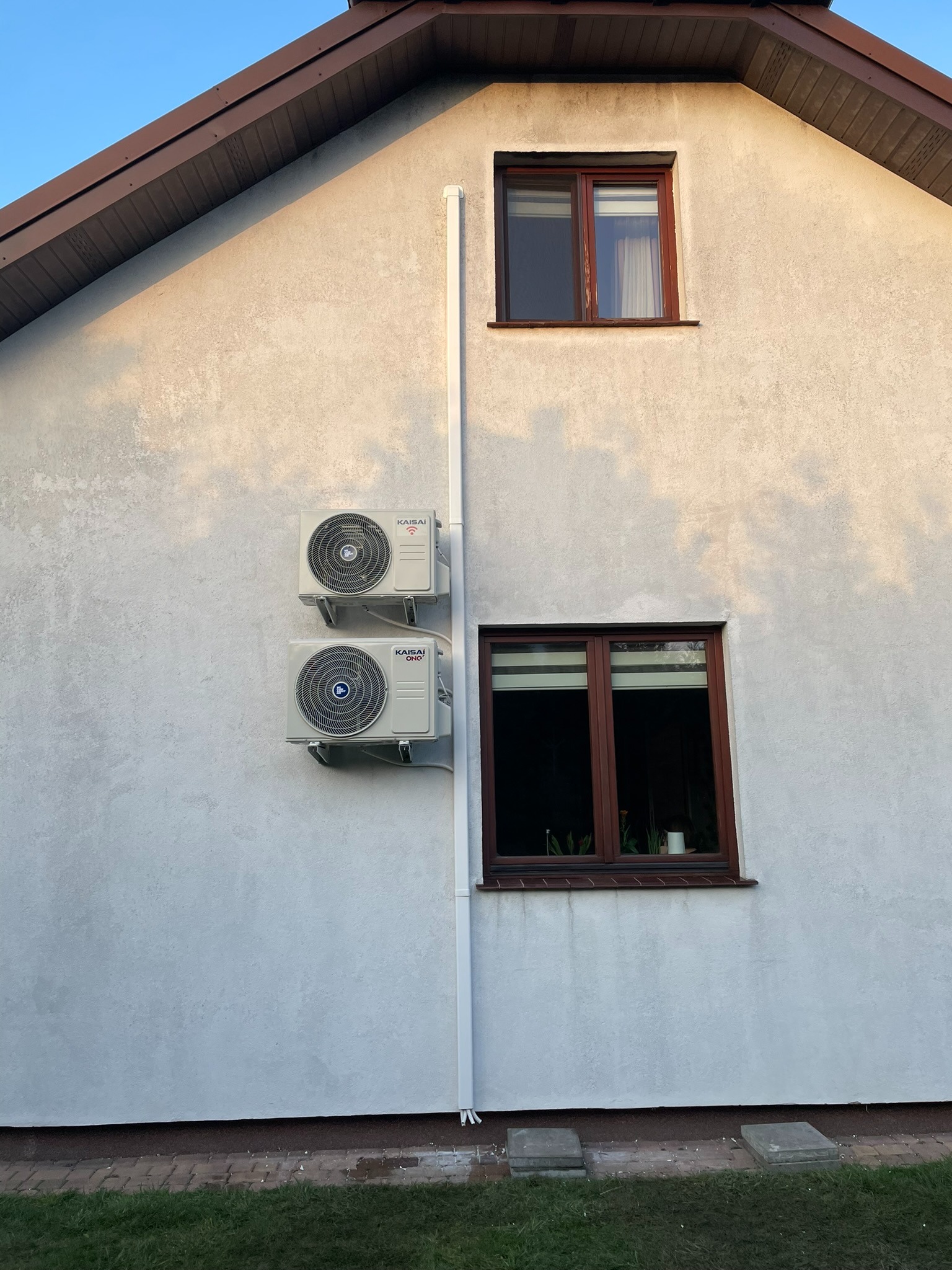 Dwa klimatyzatory marki Kaisai zamontowane na elewacji domu, widoczne okna z brązowymi ramami, biała rynna pionowa, jasna elewacja z delikatną fakturą.