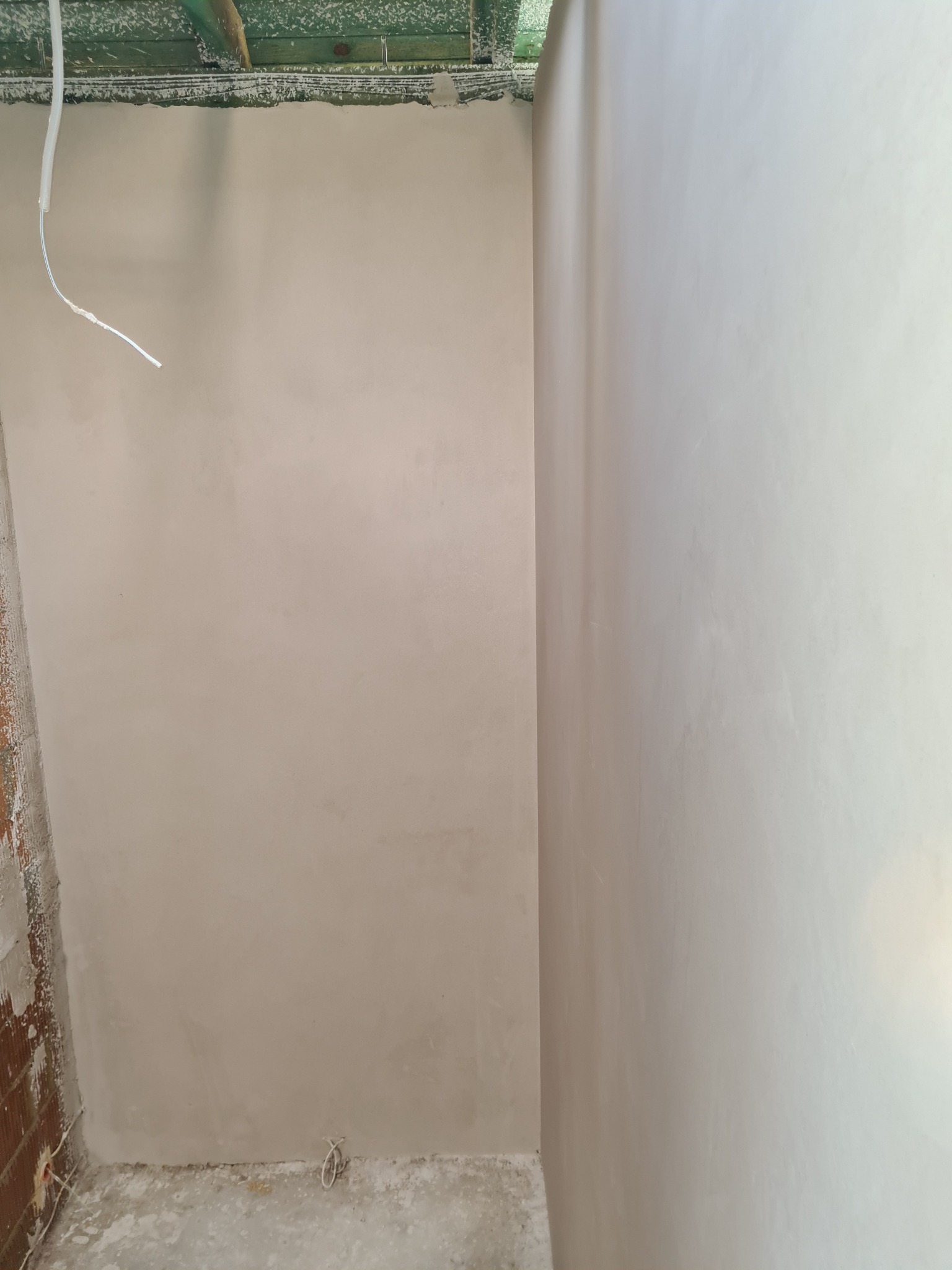 Świeżo nałożony, jasny tynk na ścianach w rogu pomieszczenia, widoczne ślady prac murarskich na sąsiedniej ścianie z cegły oraz wystający kabel elektryczny.