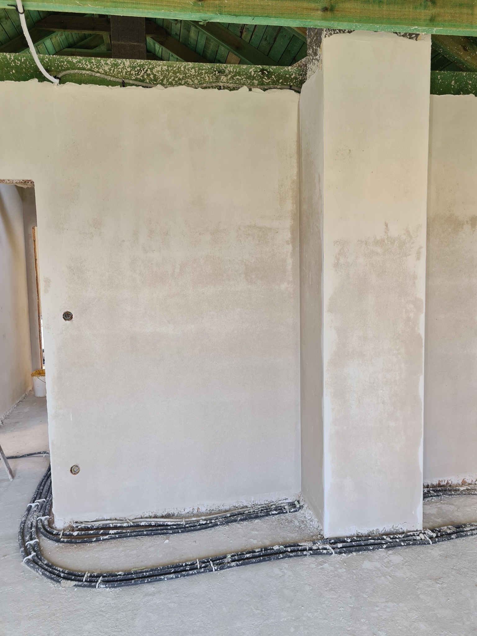 Ściana wewnętrzna po nałożeniu tynku, widoczne przewody elektryczne w peszlach ułożone przy podłodze, surowy strop z zieloną impregnacją.