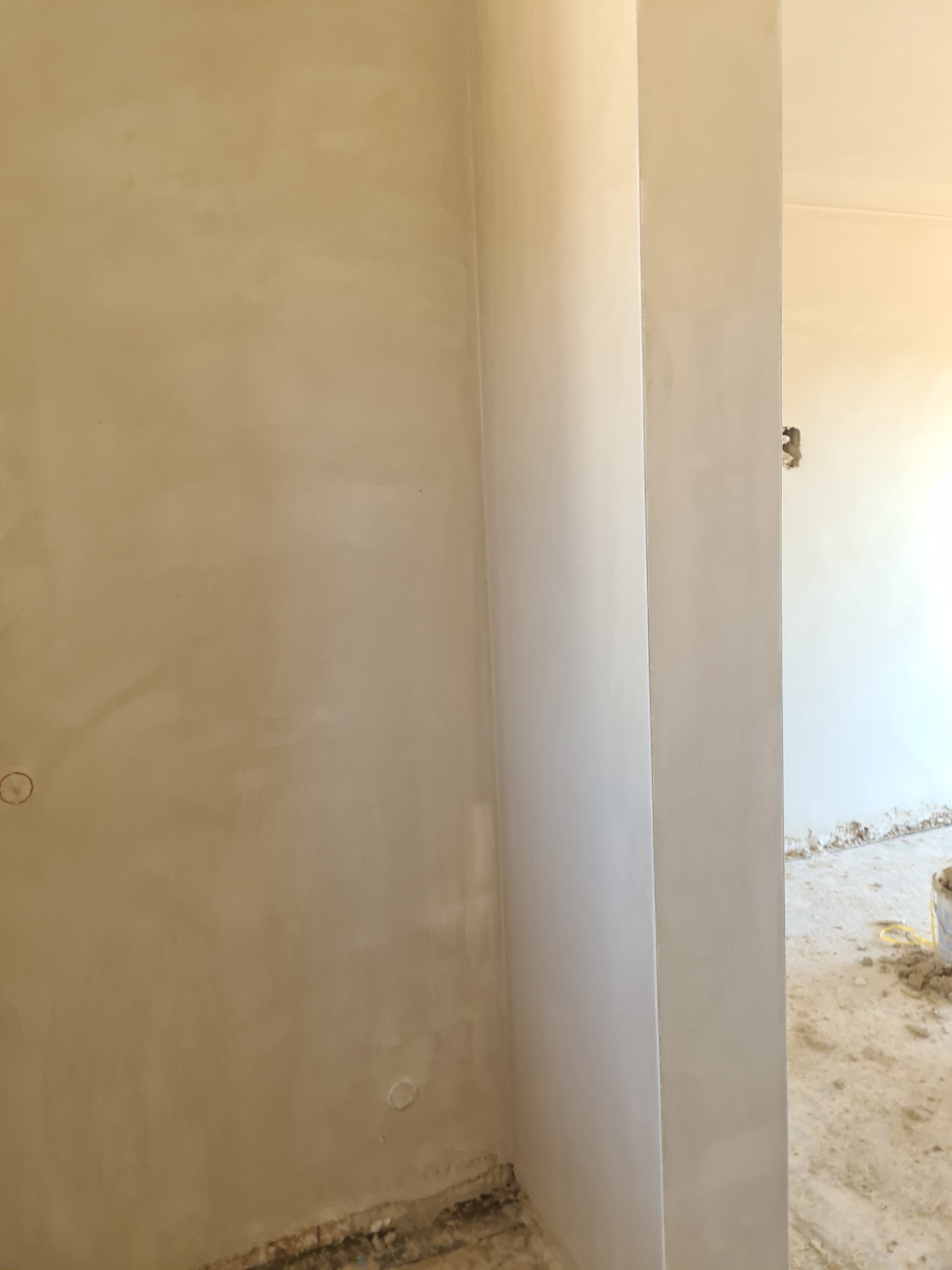 Świeżo nałożony, jasny tynk na ścianie wewnętrznej, widoczne niedoskonałości przy podłodze i w otworze drzwiowym, przygotowane do dalszych prac wykończeniowych.