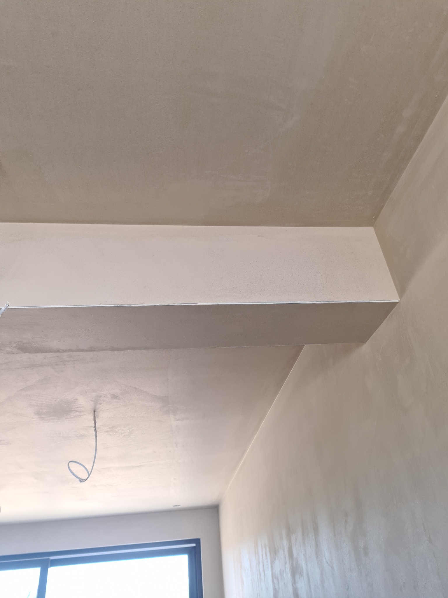 Świeżo nałożone tynki maszynowe na suficie i ścianach, widoczny wystający element konstrukcyjny z równo nałożonym tynkiem oraz wystający kabel elektryczny.
