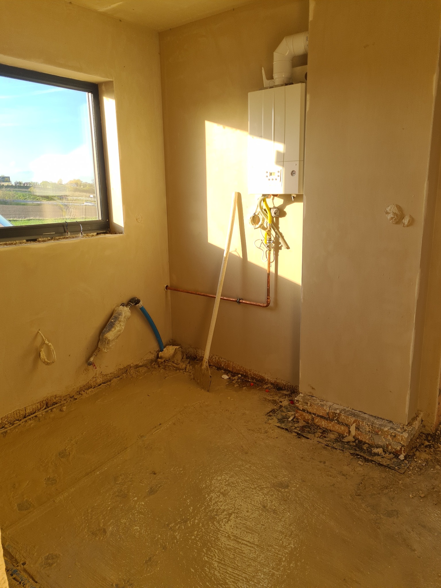 Wnętrze pomieszczenia po nałożeniu mokrego tynku maszynowego na ściany i posadzkę, widoczny piec gazowy, instalacja wodna i robocza łopata oparta o ścianę.