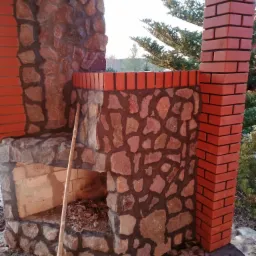 Fragment murowanego grilla ogrodowego z elementami z kamienia i cegły klinkierowej w Szczytnie, z widocznymi śladami po budowie i opartym kijem.