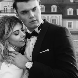 pawelzalecki.pl - Czarno-białe zdjęcie narzeczonej w białej sukni i narzeczonego w smokingu z muchą, obejmujących się na tle pałacu.