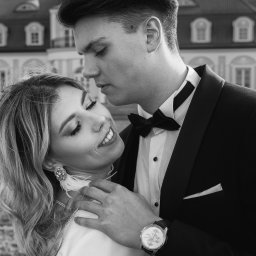 pawelzalecki.pl - Czarno-białe zdjęcie obejmującej się pary: mężczyzna w smokingu z muchą i zegarkiem na ręku oraz uśmiechnięta kobieta z ozdobnymi kolczykami na tle budynku.