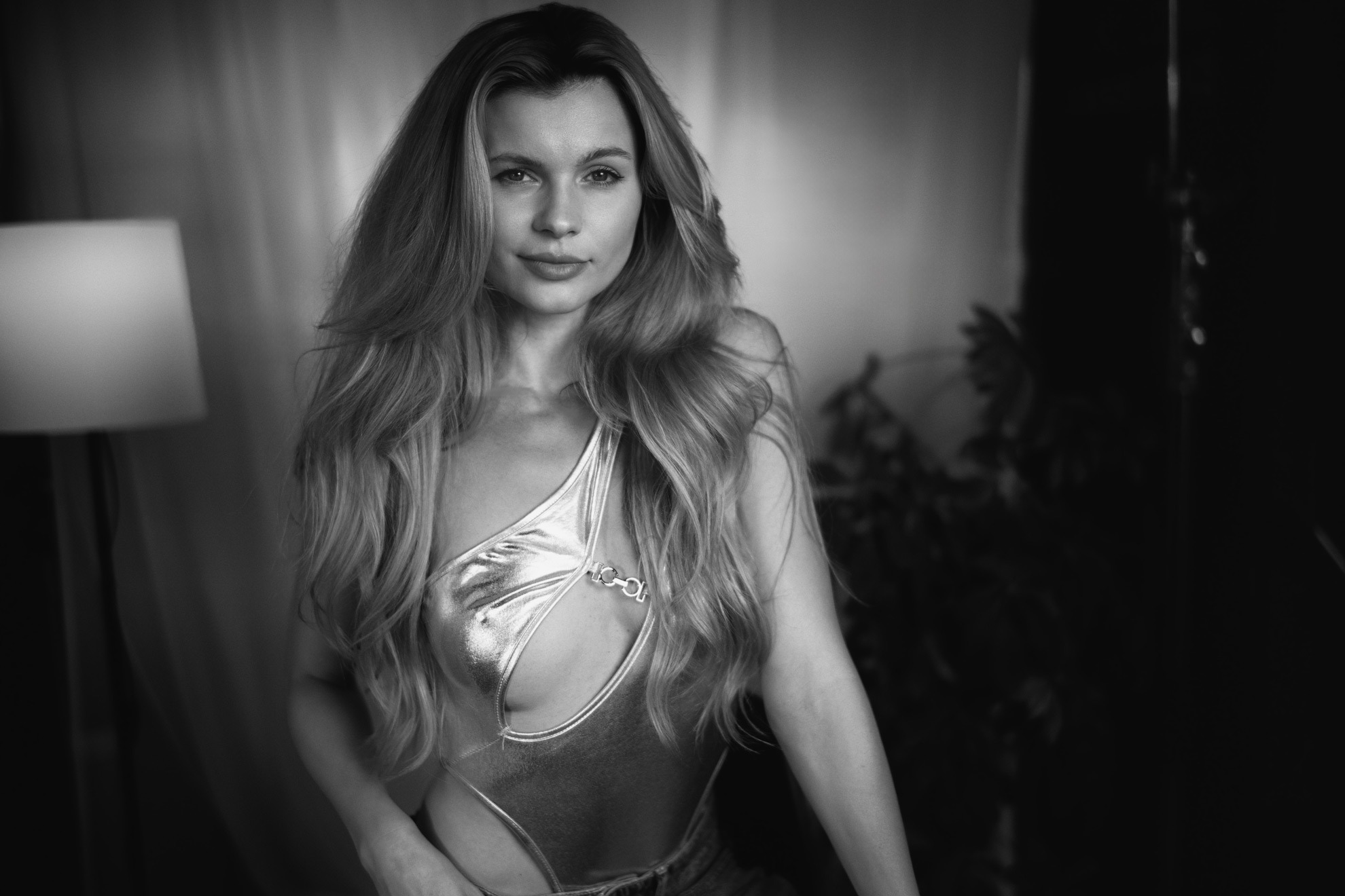 Czarno-białe zdjęcie kobiety w połyskującym stroju z wycięciem, pozującej w pomieszczeniu z lampą i zasłonami w tle.