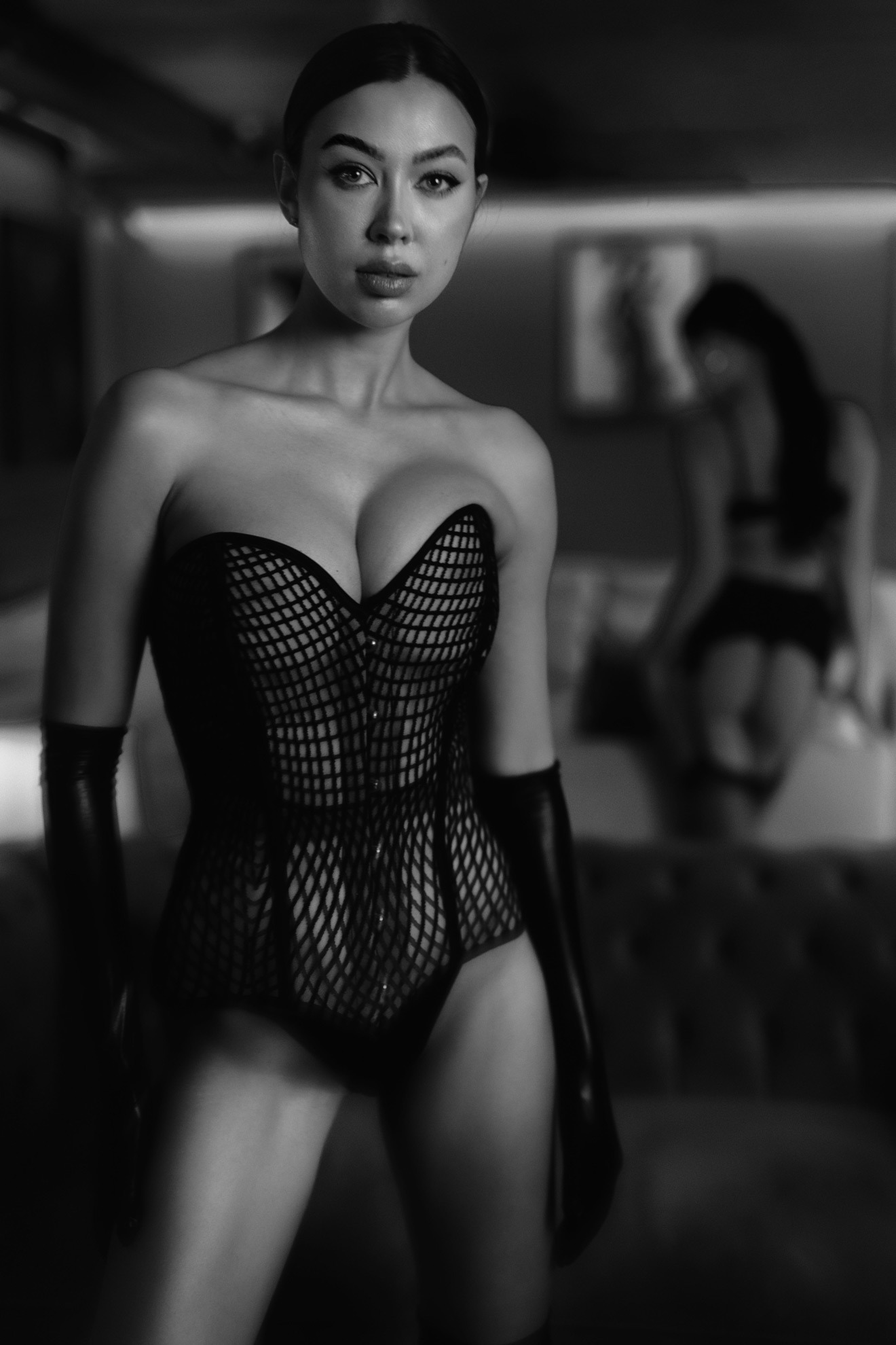 Czarno-białe portretowe ujęcie kobiety w siatkowym gorsecie i długich, błyszczących rękawicach, z drugą, zamazaną postacią w tle siedzącą na sofie.