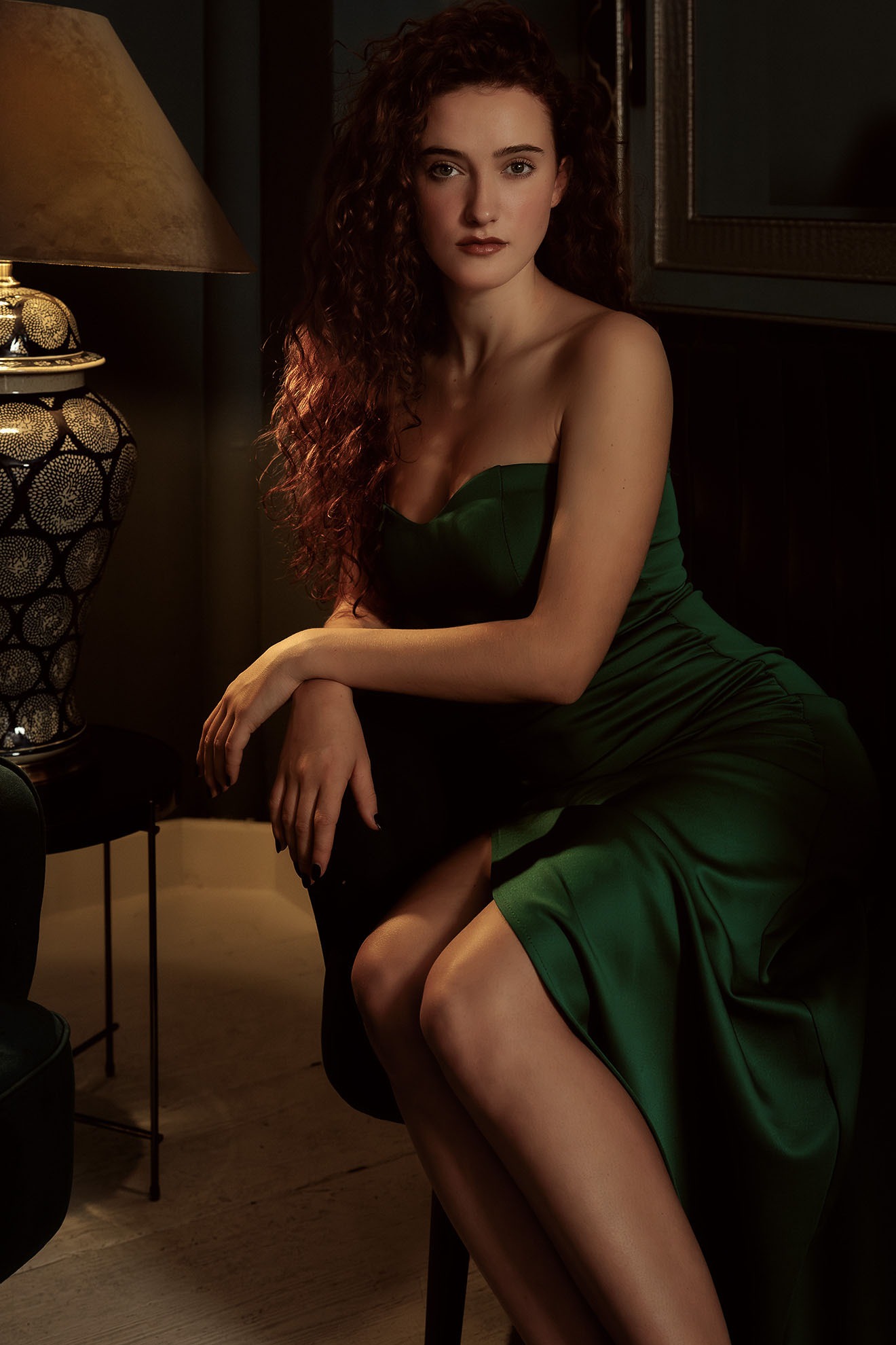 Portret kobiety w szmaragdowej sukni, z kręconymi włosami, pozującej w eleganckim wnętrzu w Warszawie, z dekoracyjną lampą w tle.