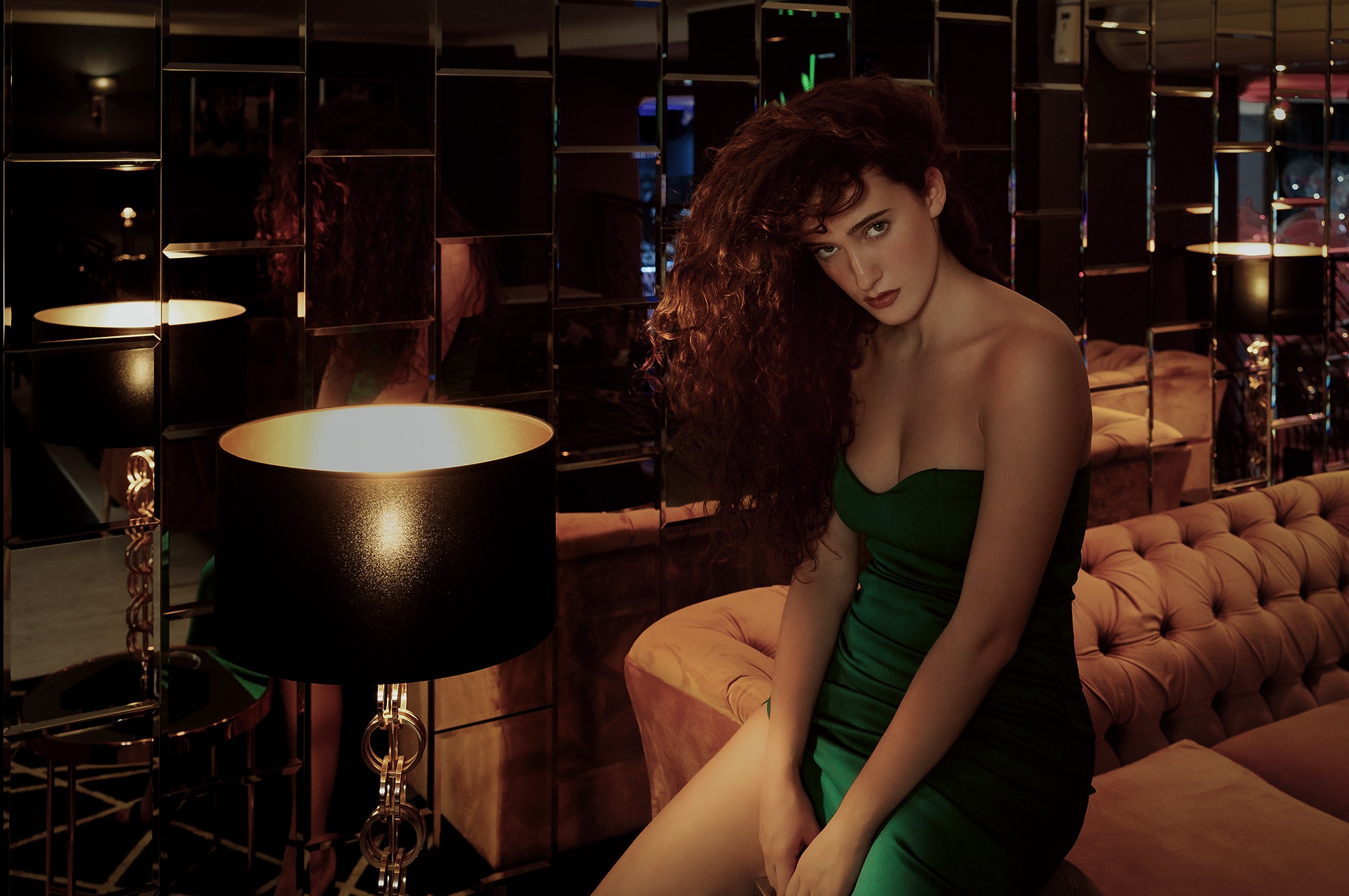 Portret kobiety w zielonej sukience bez ramiączek, siedzącej na pluszowej kanapie z lampą z czarnym abażurem i złotymi detalami, na tle ściany z lustrzanych płytek.