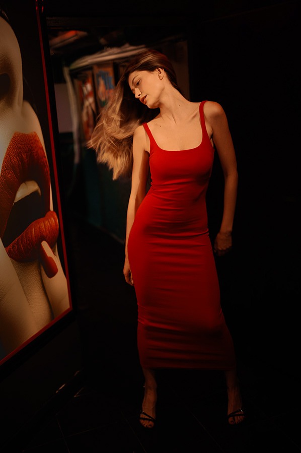 Kobieta w obcisłej czerwonej sukience pozuje na tle ciemnego pomieszczenia z plakatem przedstawiającym usta.