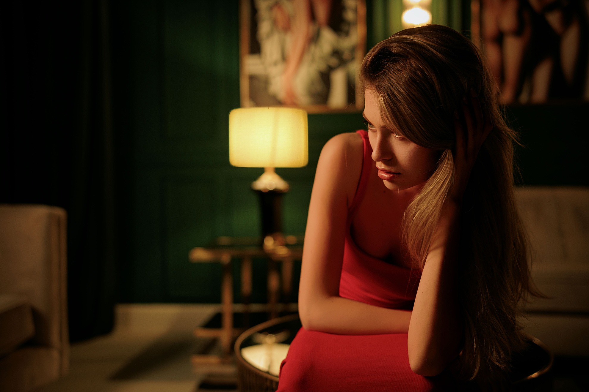 Zamyślona kobieta w czerwonej sukience siedzi w pokoju z zielonymi ścianami, oświetlonym ciepłym światłem lampy, z ręką we włosach i delikatnym wyrazem twarzy.