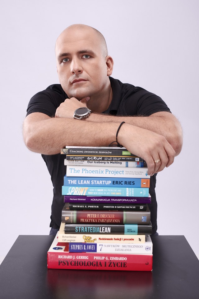 Portret mężczyzny w czarnej koszulce opierającego się na stosie książek o tematyce biznesowej i rozwoju osobistego, widoczny zegarek na nadgarstku i obrączka na palcu.