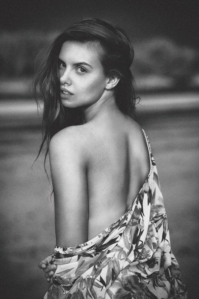 Czarno-białe portretowe ujęcie kobiety z odsłoniętymi plecami, spoglądającej przez ramię, okrytej kwiecistą tkaniną.