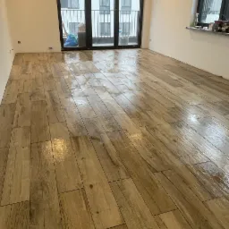 Świeżo ułożona, błyszcząca podłoga z paneli imitujących drewno w jasnym, pustym pokoju z balkonem i oknem; widoczne odbicia światła na powierzchni.