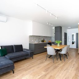 M2 Visual - Jasne wnętrze: szary narożnik, drewniana podłoga, stół z kwiatami, kuchnia z szarymi szafkami i marmurowym blatem, białe ściany, designerskie lampy sufitowe.