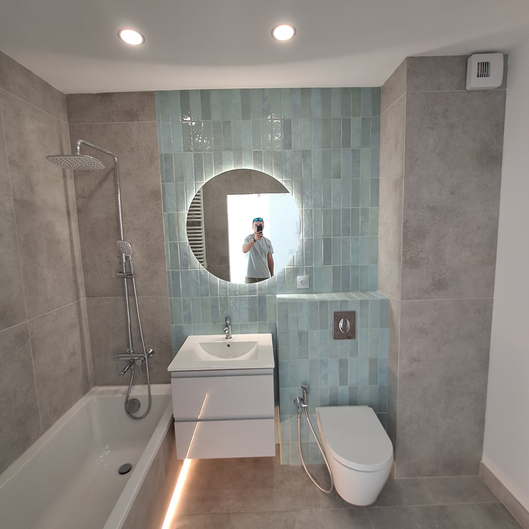 Nowoczesna łazienka w szaro-błękitnej kolorystyce z podświetlaną umywalką, okrągłym lustrem z podświetleniem LED i widocznym odbiciem fotografa w lustrze.