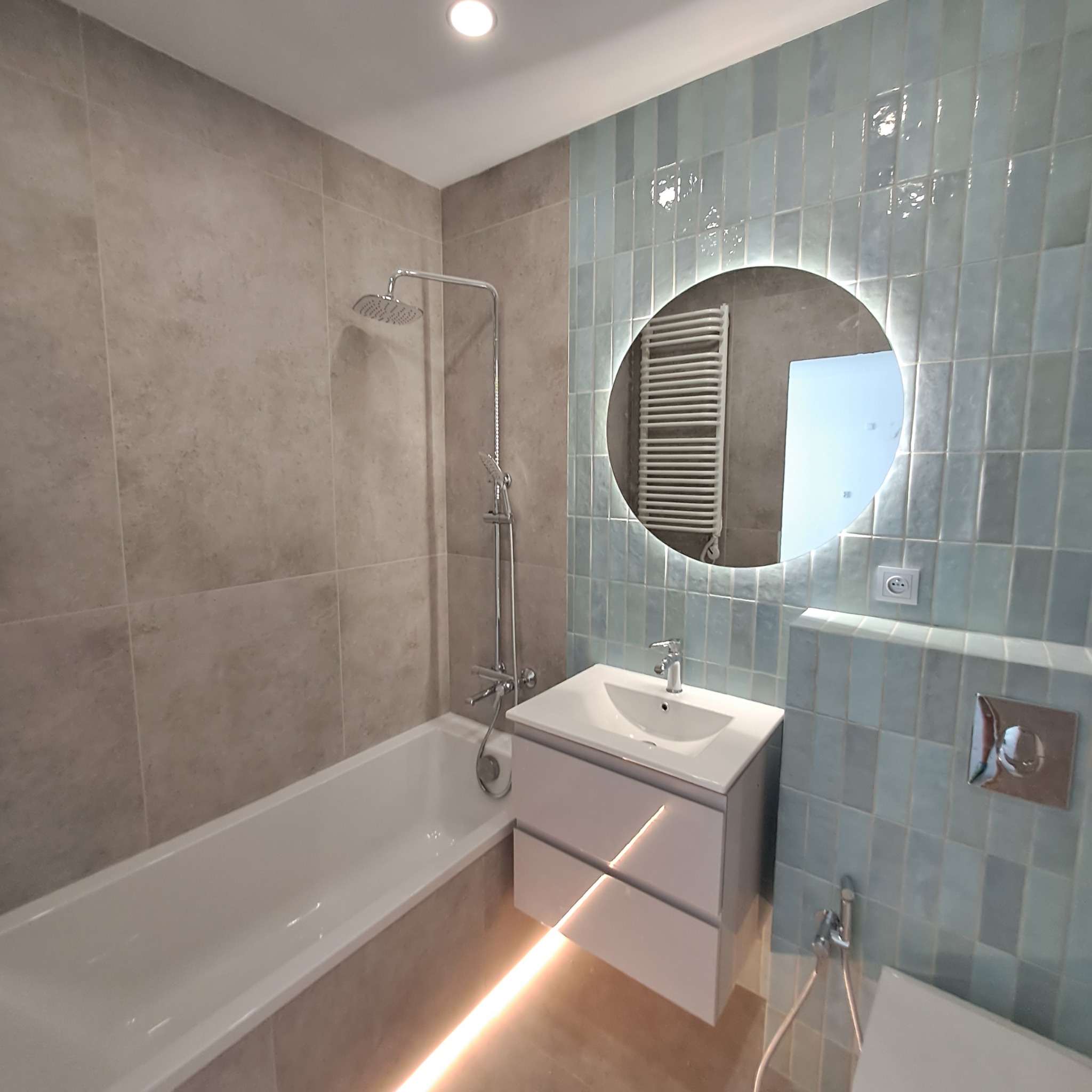 Nowoczesna łazienka po remoncie: wanna z chromowaną armaturą, umywalka z szafką podświetloną LED, lustro z okrągłym podświetleniem, płytki w odcieniach szarości i błękitu.